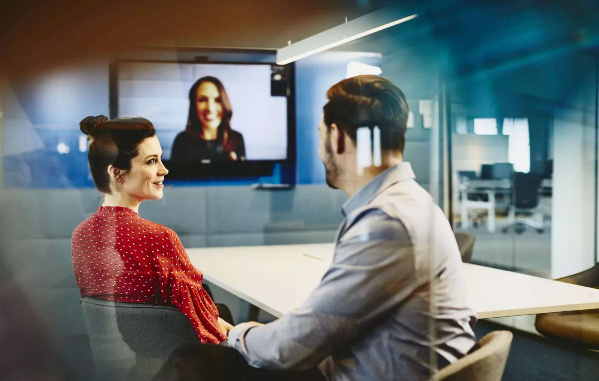 2 mensen zijn aan het vergaderen in een zaal samen met een 3e persoon op het videoscherm. Inclusieve communicatie waarbij iedereen zich betrokken voelt.